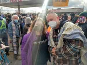 Two people in masks amid a large crowd smile at the camera. One wears a shirt that says "Jews for Black Lives" and the other wears a tallit and a kippah