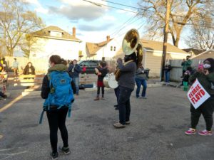 A spaced out circle of people play instruments including tuba, drum, tambourine, and trumpet. Other people look on and take photos and video on their phones.
