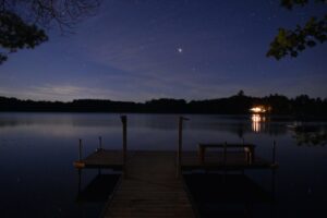 Dusk over a lake, with the photo taken overlooking a dock and with the other side of the lake with trees visible. Blue and purple hues across the image.