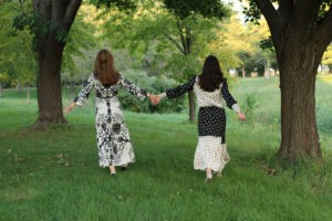 Two women with long hair and dresses running away from the camera holding hands.