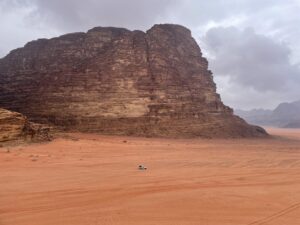 Photo of a tall rock structure in the desert with a car driving in front of it, though the photo is taken from such a distance that the car is tiny compared to the giant rock.
