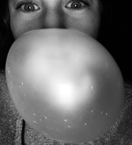 A person looking at a camera in a close up black and white image with the person blowing a balloon.
