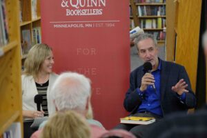 Author Sam Freedman (right) in a blue shirt and black sports jacket, gestures with his hand to an audience while Star Tribune editor Laura McCallum (left) smiles at a book event for "Into the Bright Sunshine" at Magers & Quinn Booksellers in Minneapolis