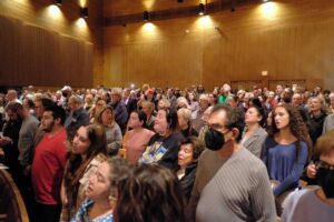 Hundreds of people stand in a synagogue sanctuary that has warm tones and lighting. The people have their mouths open because they are singing.