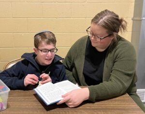 A woman, at right, wearing a green sweater and a ponytail, reaches over to look at a book that a boy in a kippah and glasses, at left, is reading from.