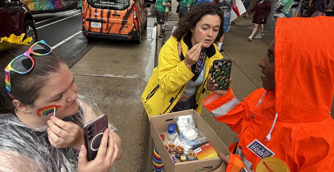 Milo Beyene (right) holds their phone up for Molly Knopf to put glittter on her face. At right Sarah from Sequeerity puts glitter on.